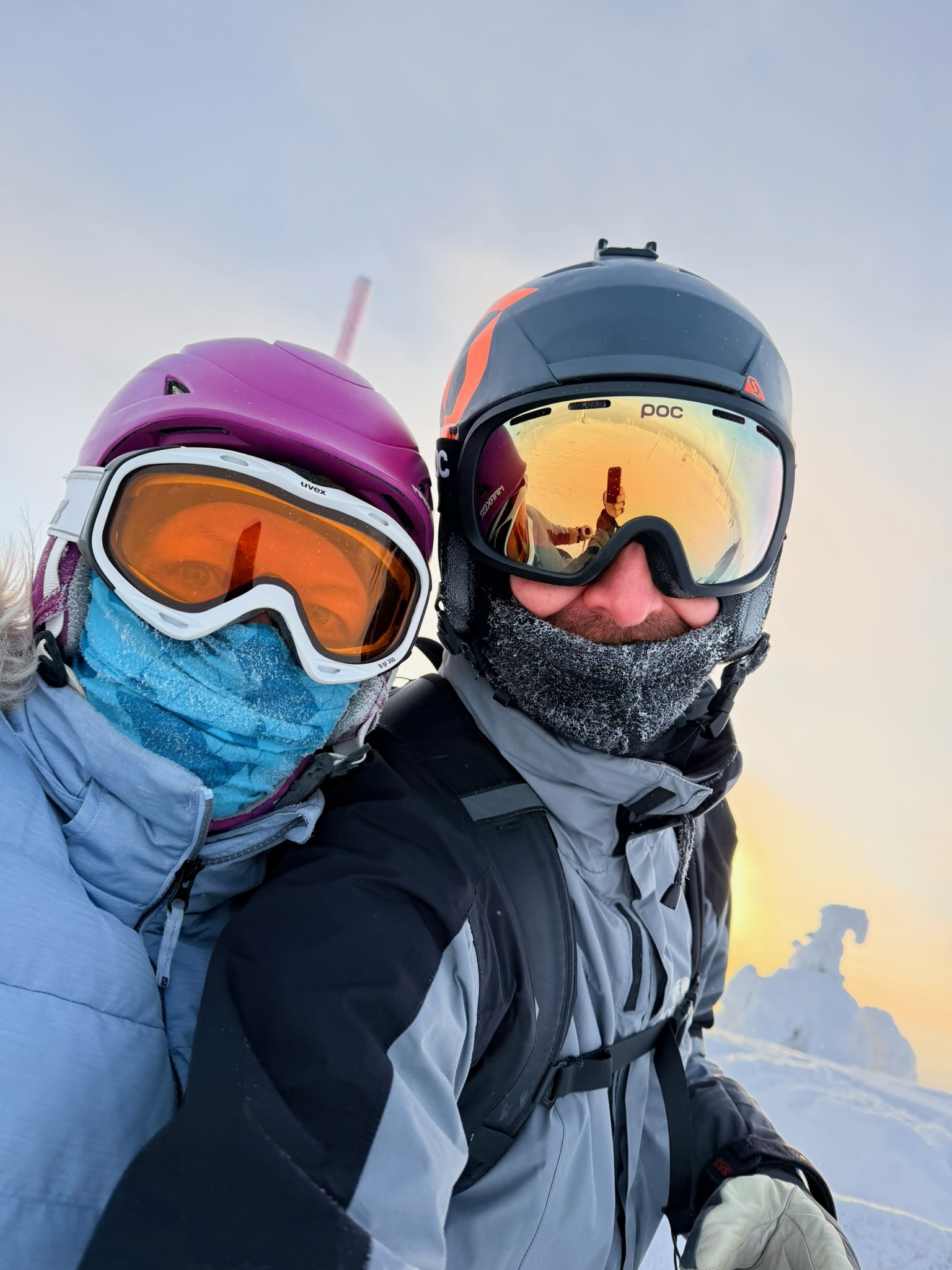 Couple selfie at the summit — snow ghosts behind us, sun just over the treeline